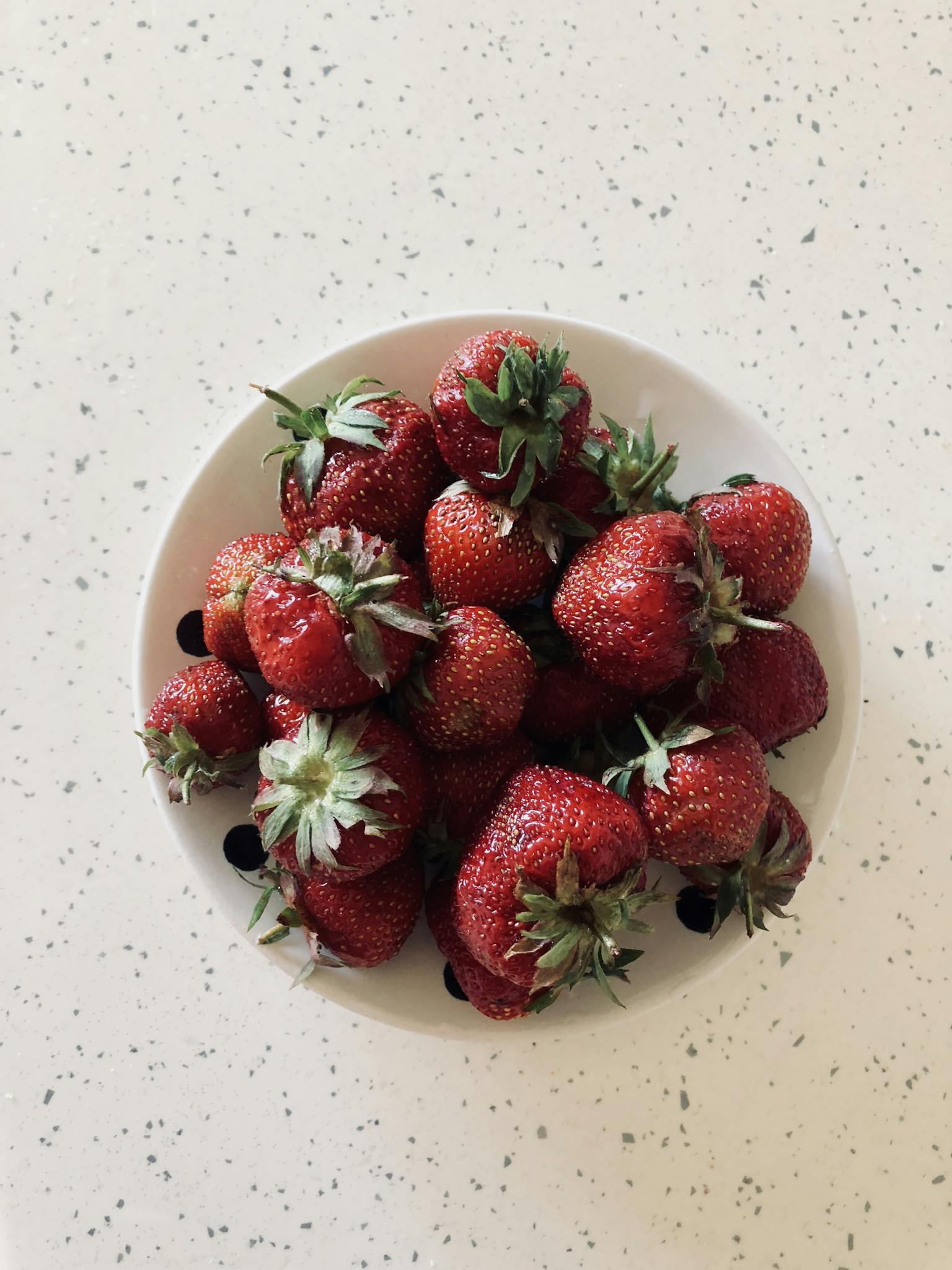 Aerial view of fresh strawberries in a white bowl on a speckled countertop.