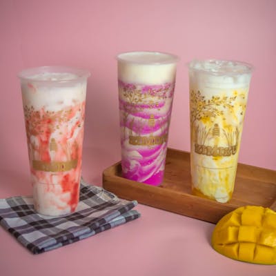 Plastic glasses of tasty cold milkshakes of different flavors served on pink surface near ripe juicy mango