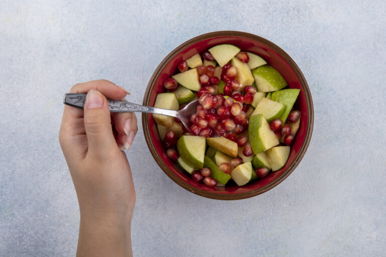 Top view of female hand holding spoon with pomegranate seed and chopped apples in a red bowl on white background